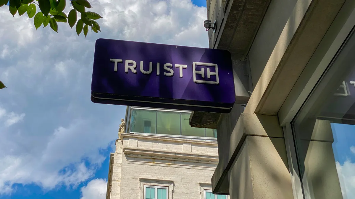 A purple "Truist" bank sign attached to the facade of a stone building is shown, with blue sky and a tall building behind it.