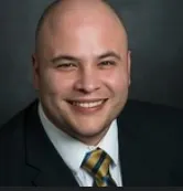 A headshot of a man smiling at camera in a suit with a striped gold necktie.