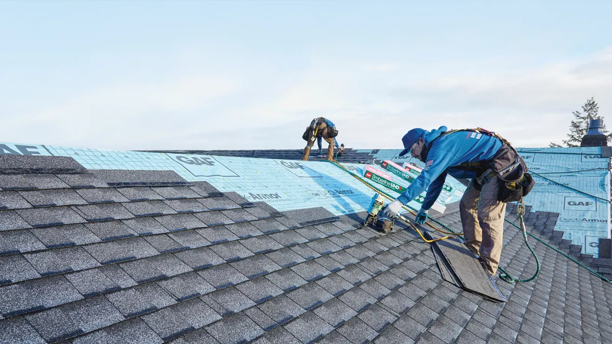 Two workers in safety gear install shingles on a gray roof with blue underlayment, under a clear sky, conveying teamwork and focus.