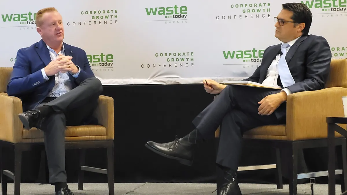 Two people sit in chairs on a stage with a white background behind them that says "Corporate Growth Conference" in green letters.