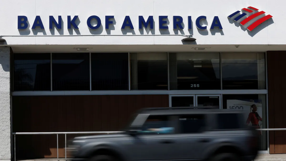 A car drives by a brick and glass storefront with the words "Bank of America" and the Bank of America flag logo above the entrance.