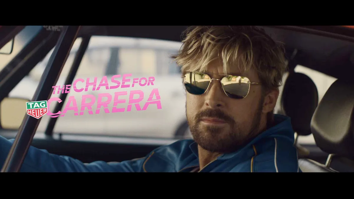 Ryan Gosling holds a steering wheel while wearing sunglasses and a blue tracksuit.