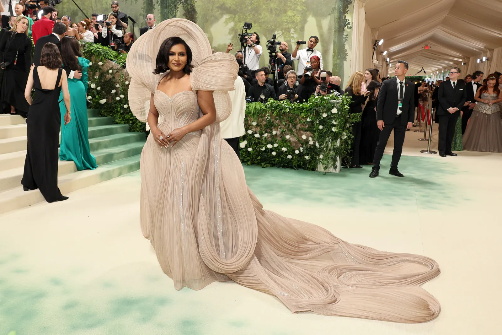 A person in a beige dress with looped embellishments stands in front of photographers.