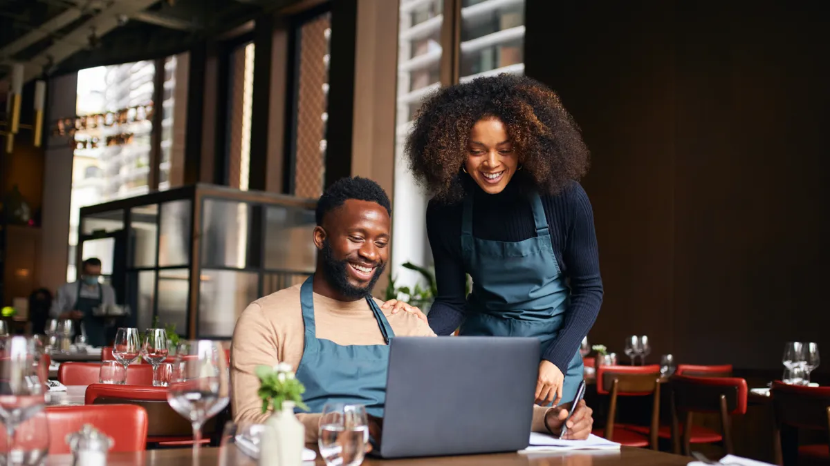 Two restaurant workers at a table smiling and looking at a laptop.