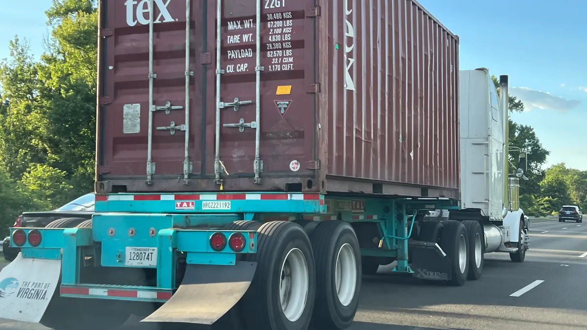 A truck hauls a red Textainer shipping container on a blue chassis with Port of Virginia mudflaps on tree-lined Interstate 95 North in Maryland.