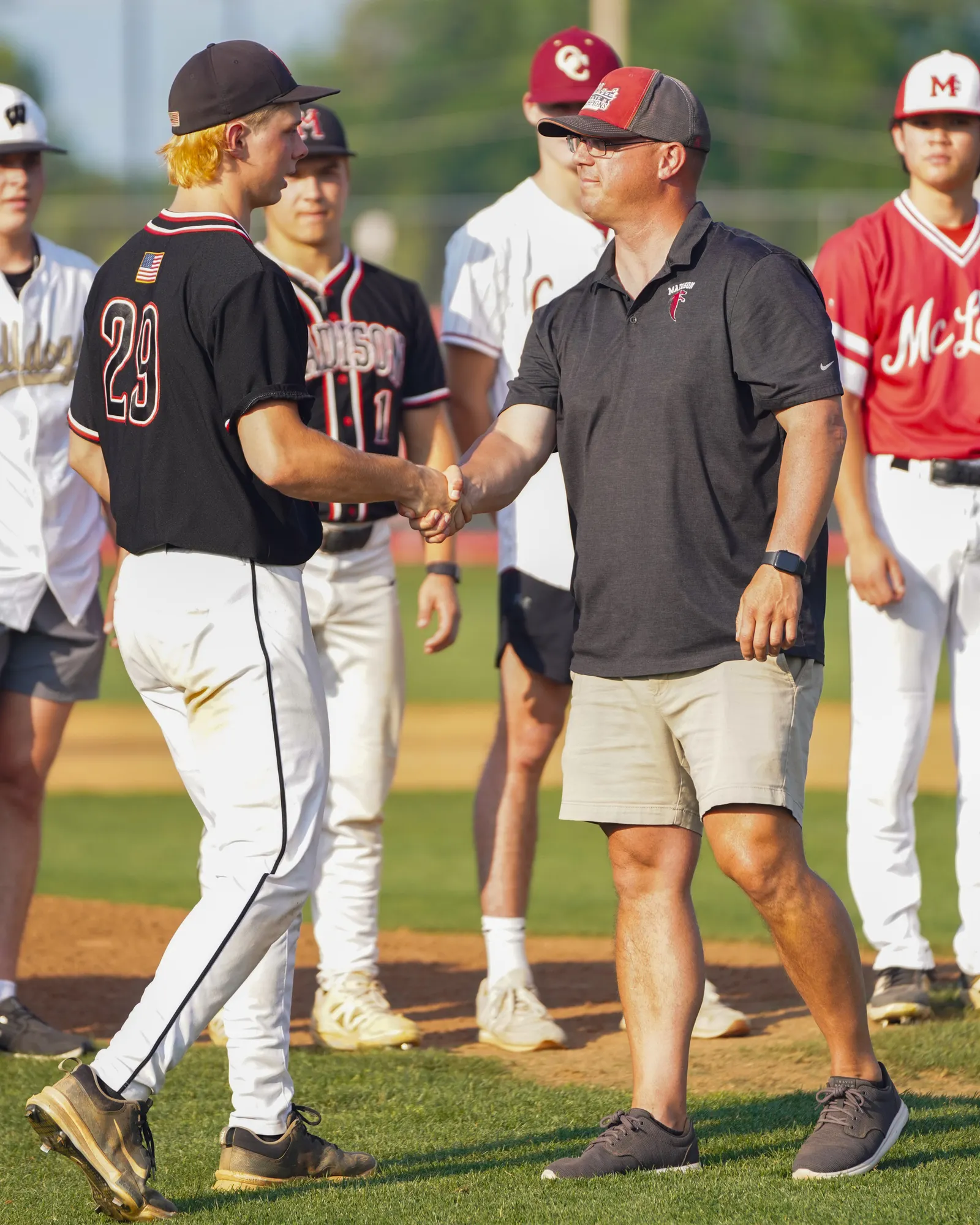 An adult is shaking hadns with a youth baseball player outside near a field. Other baseball players are nearby