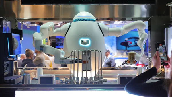 A white oval-shaped robot with arms holds two drinks inside a modern mini kitchen; a person with a phone takes a picture of the robot on the right.