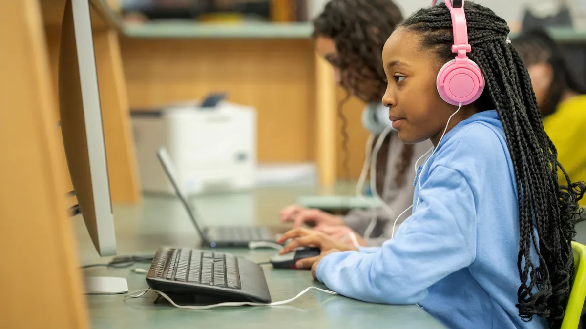 A student wearing pink headphones looks at a desktop computer in a school library computer lab, surrounded by other students working on laptops.