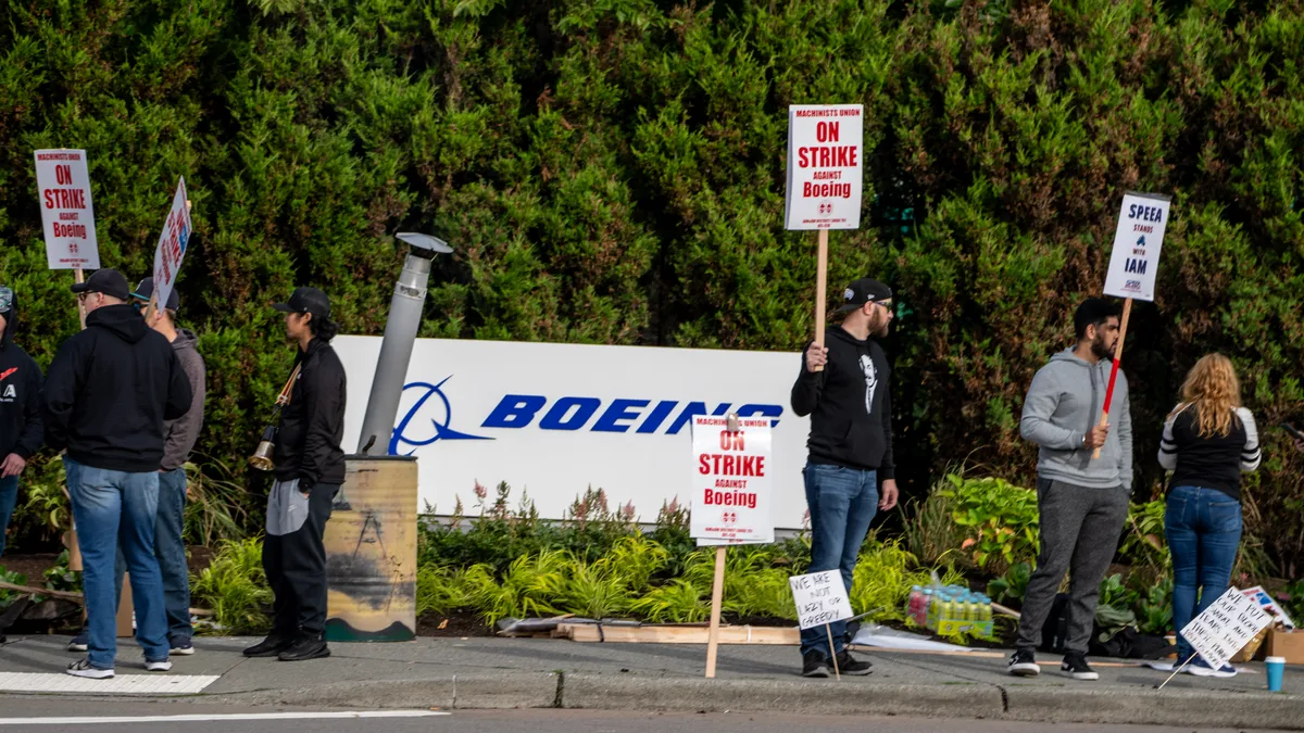 A group of eight people holding red and white picket signs in front of a blue and white Boeing sign.