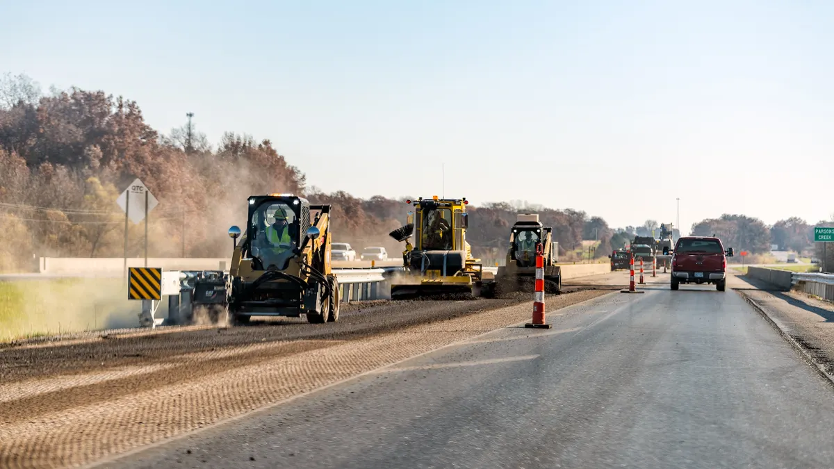 Several construction vehicles on an unpaved section of a highway with cars passing on the adjacent pavement.