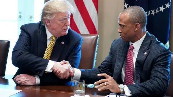 Two seated people in suits turn to one another and shake hands.