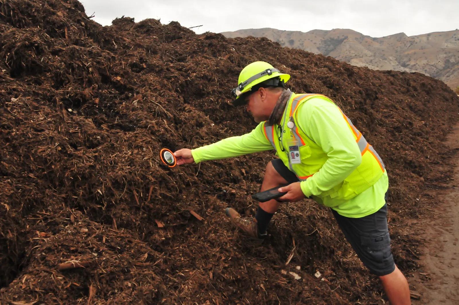 Man in safety gear and hat checks the temperature of a compost pile