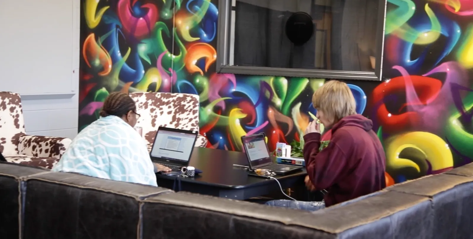 Two people sit on a couch. Open laptops are sitting on a table in front of them. A colorful design is shown on a wall in front of them.