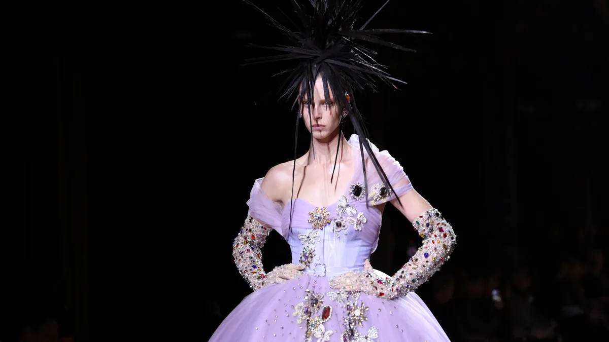 A model wearing a black spiked headdress, jeweled gloves and a full-skirted lavender dress walks down a darkened runway.