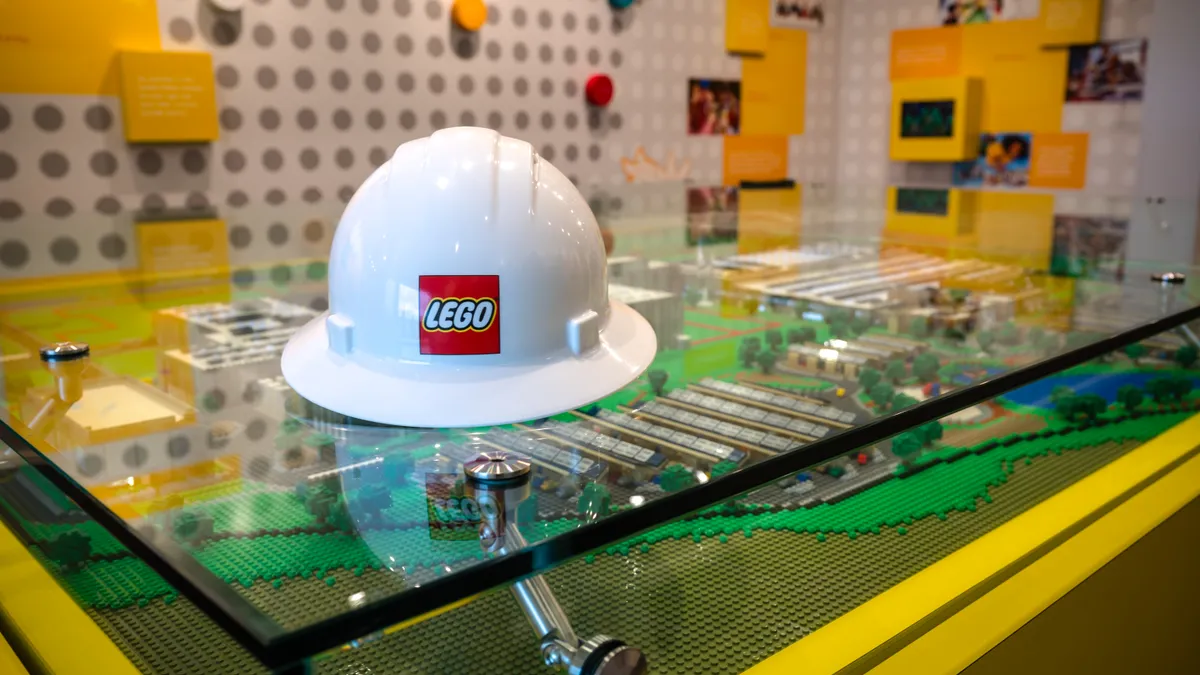 A white hardhat with the red Lego logo on its front, laying on glass covering a Lego rendering.