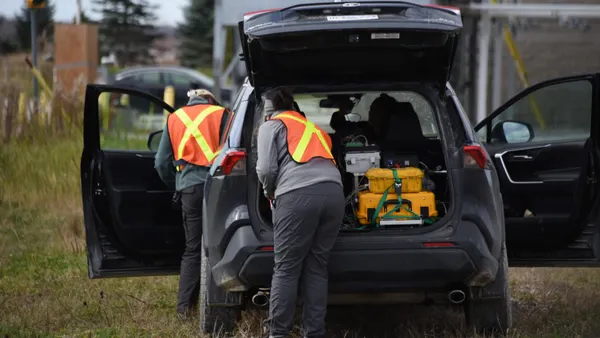 Two people wearing hi-vis construction vests stand around a sport utility vehicle. Scientific instruments are visible inside the car.