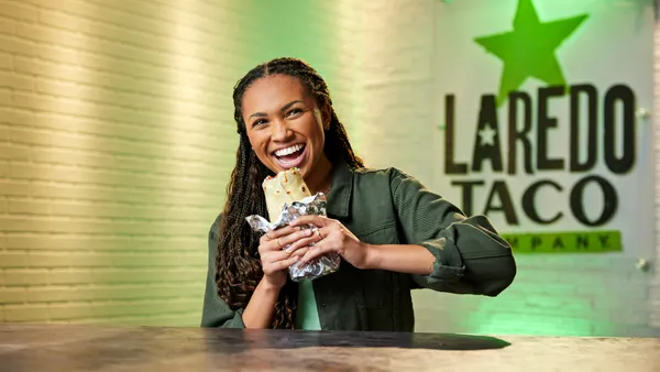 A photo of a smiling person sitting at a table, about to bite into a burrito. A sign on the wall behind them says Laredo Taco.