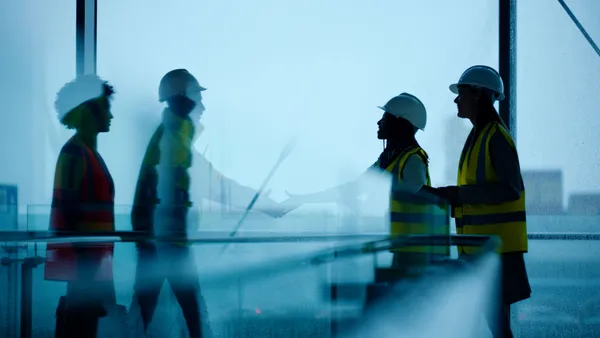 Two pairs of people in construction gear shake hands in a building on a rainy day.