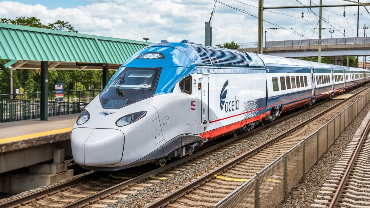 A sleek blue and white Amtrak Acela high-speed train is seen passing a station with a green canopy and overhead wires.