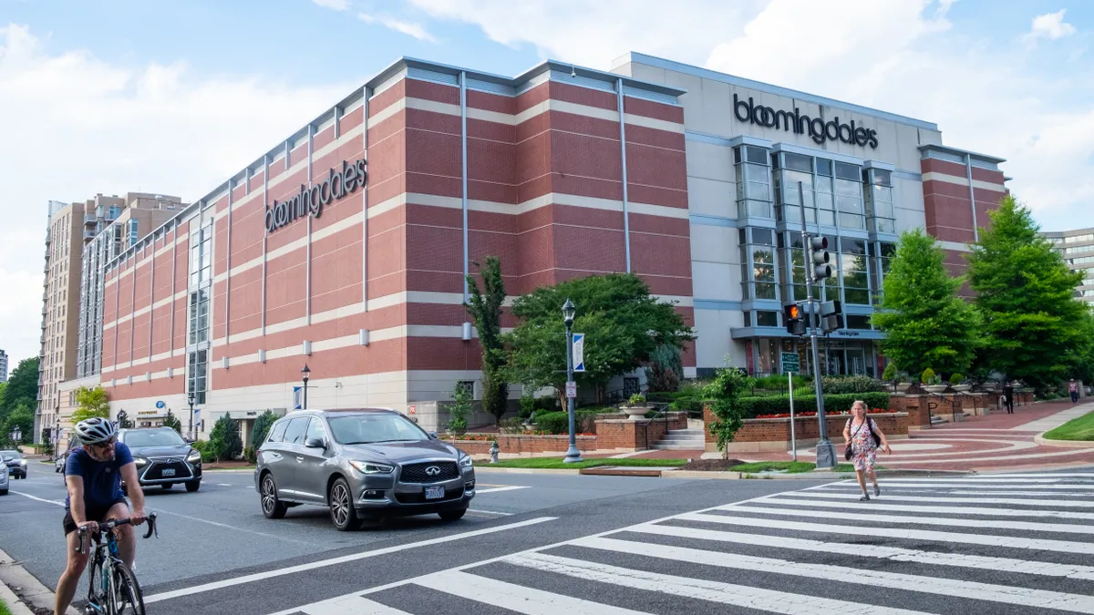 Traffic including cars and a cyclist ride by a Bloomingdale's store under blue skies, as a nearby pedestrian crosses the street.