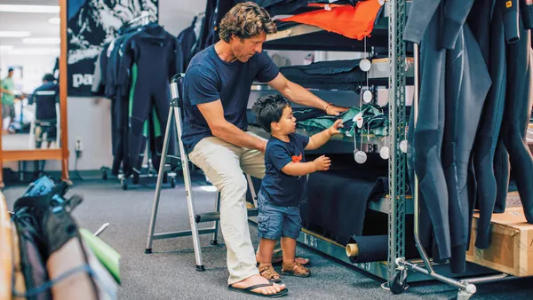 A man sitting in a chair looks at samples of wetsuit fabric with his toddler son, who is standing beside him.