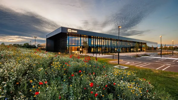 A picture of building with large glass windows and a parking lot, with flowers and grass in the foreground