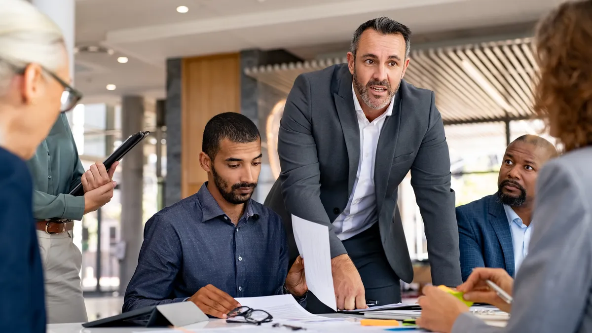 Man standing with hands on table at table at a meeting with other people in professional setting.