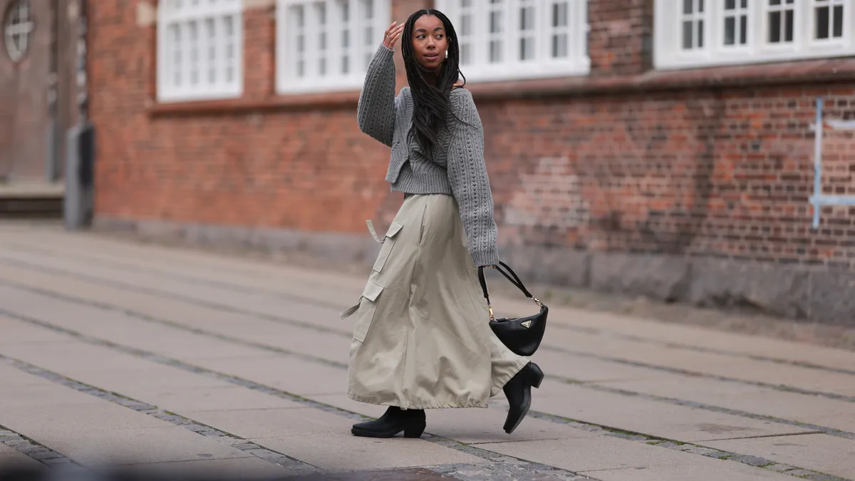 A woman walks down a wide, quiet street in Copenhagen wearing a grey wool knit pullover, long beige cargo skirt, black Prada bag and Copenhagen Studios Waxed Nabuc Black leather boots.