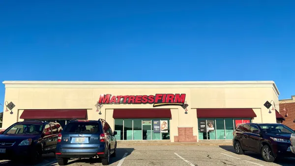 Cars are parked in front of a store with "Mattress Firm" in red capital letters, against a bright blue sky.