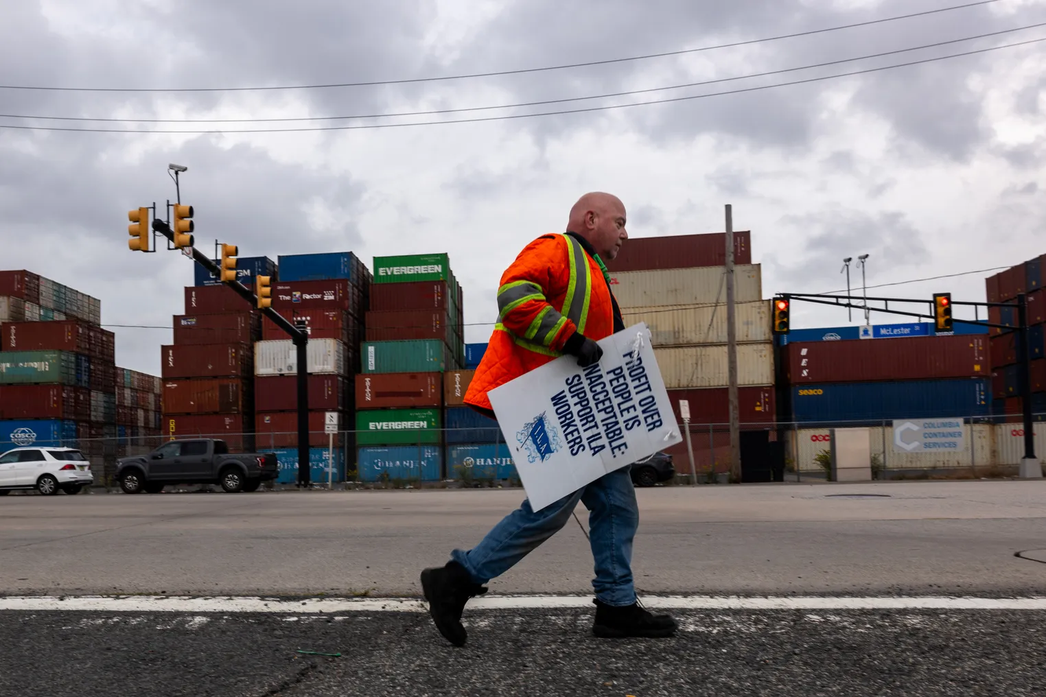 A dockworker on strike walks past shipping containers holding a sign.