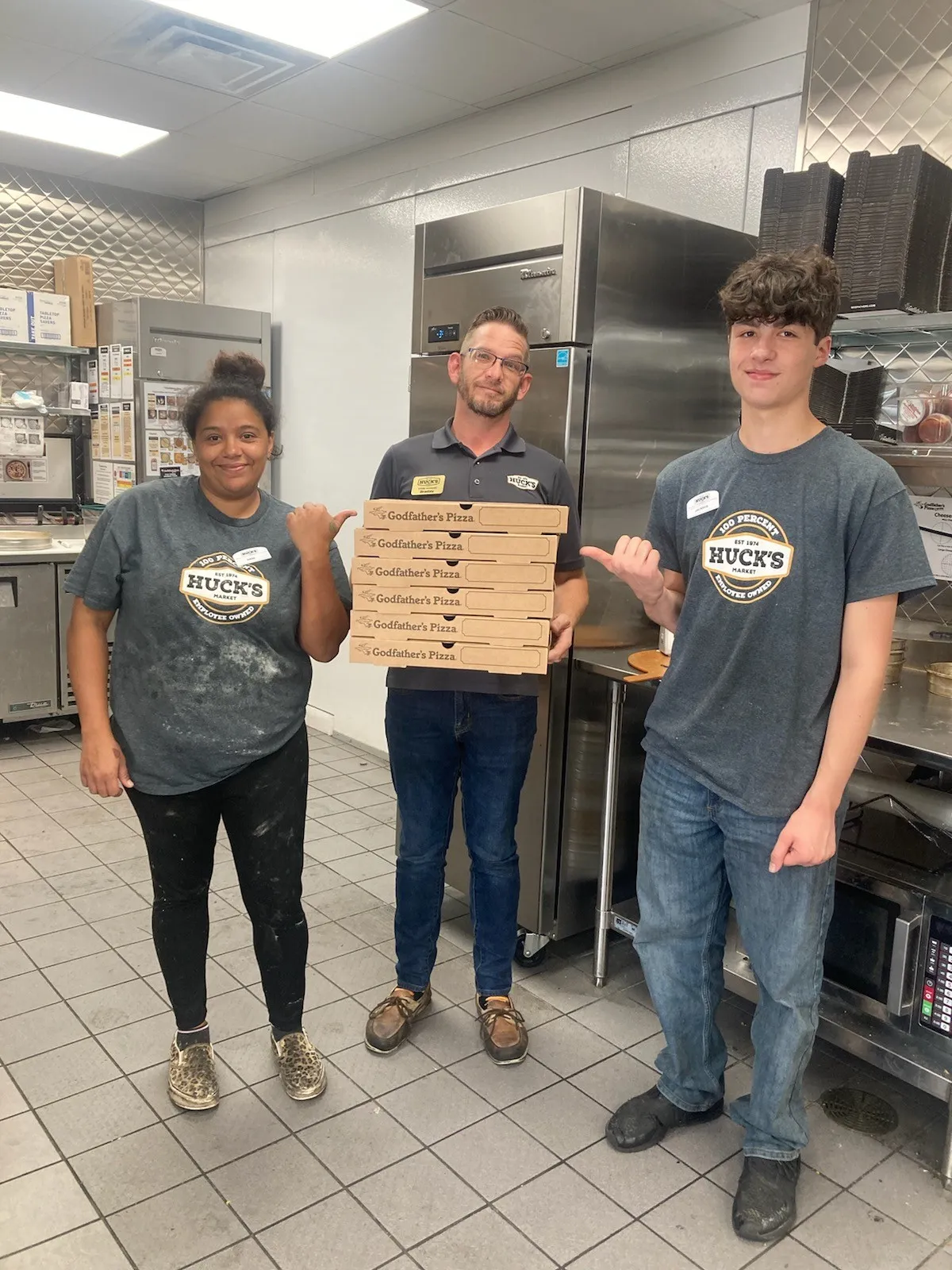 A photo of several people in the kitchen along with a stack of Huck's pizza boxes.