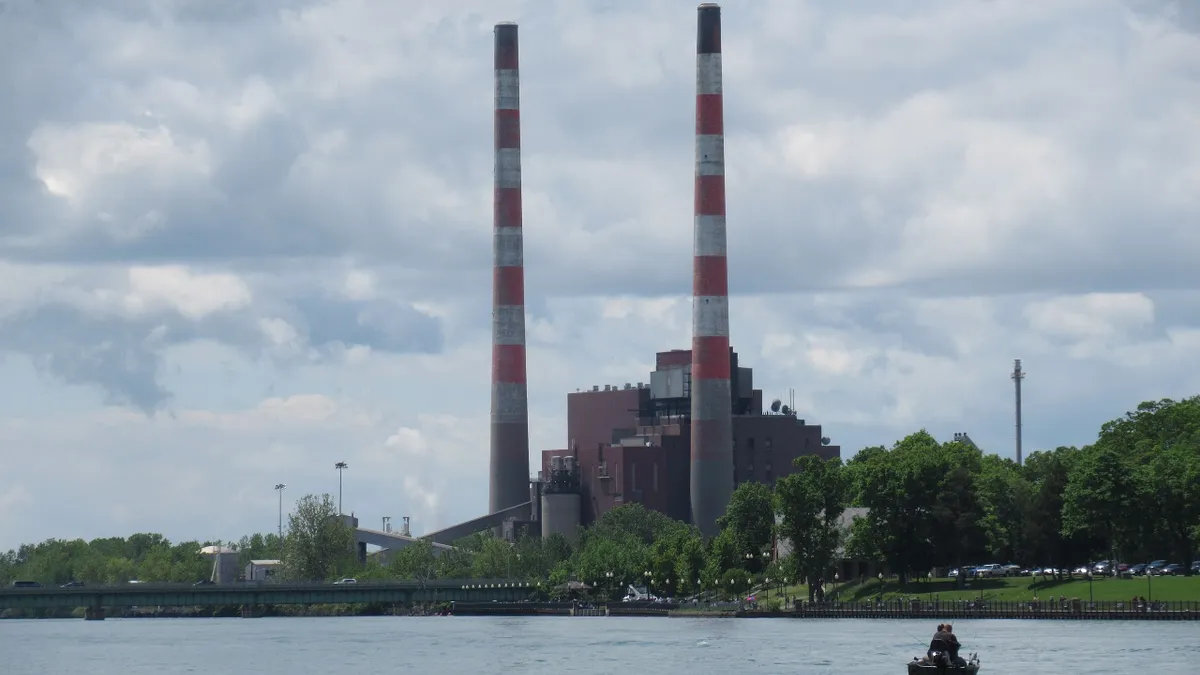 Two red- and white-striped smokestacks rise next to a large brick building next to a body of water.