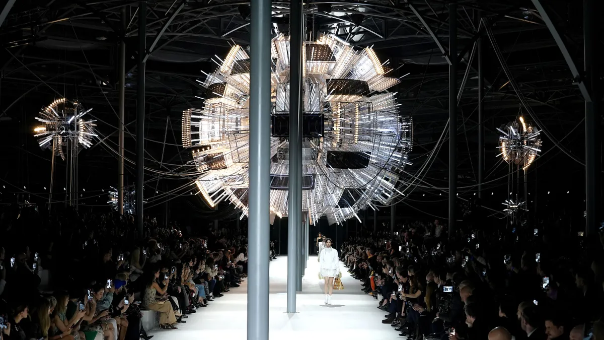 Models walk the runway beneath an elaborate light fixture.