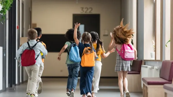 A handful of students carrying backpacks are walking or running down a school hallway. Their backs are toward the camera.