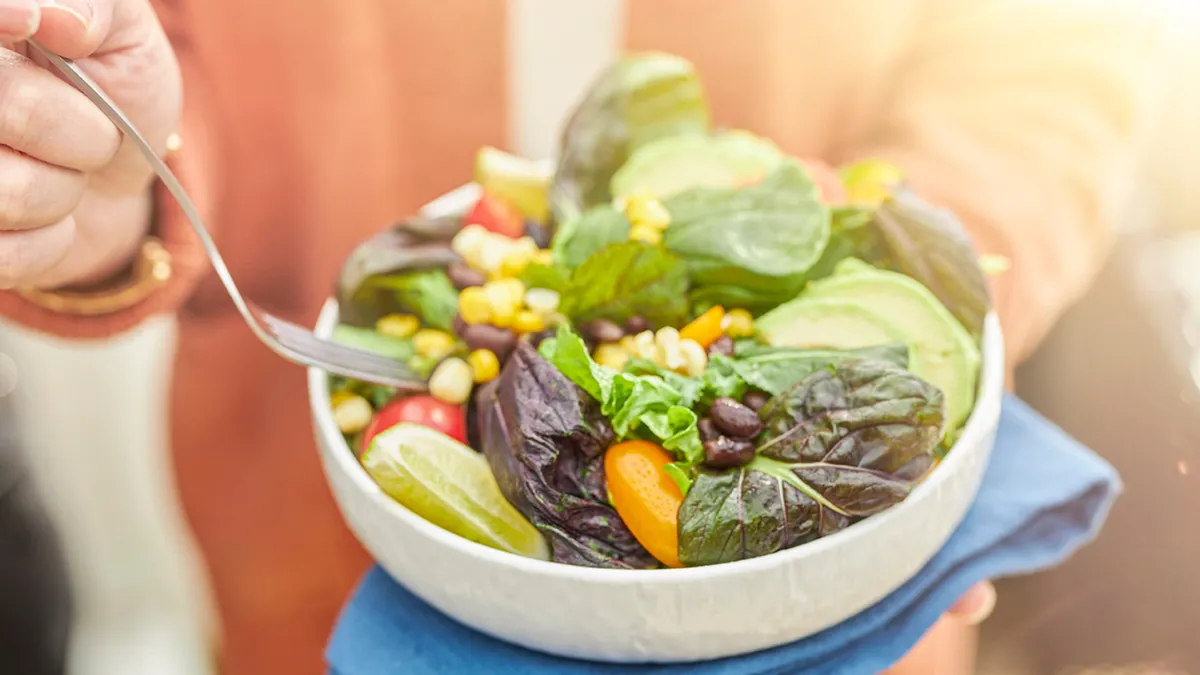 A person holds a white bowl with a salad featuring purple and green Conscious Greens, corn, beans and other vegetables.