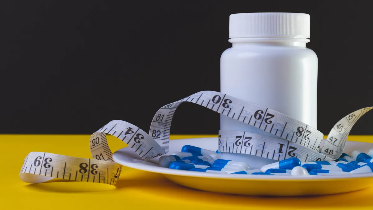 A white pill bottle, pills and a measuring tap sit on a dish on top of a yellow table