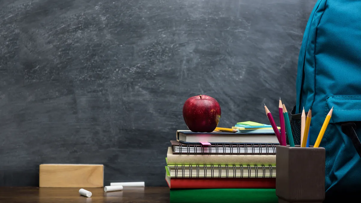 A stack of books sits on a desk alongside chalk, eraser, a pencil holder and backpack. A red apple sits on top of the stacked books.