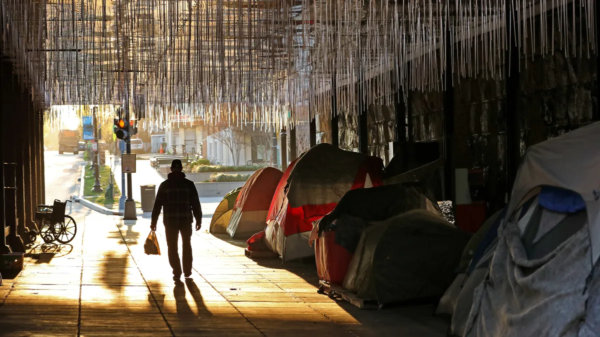 Tents lined up underneath a bridge. A person walks by, silhouetted by the sun.