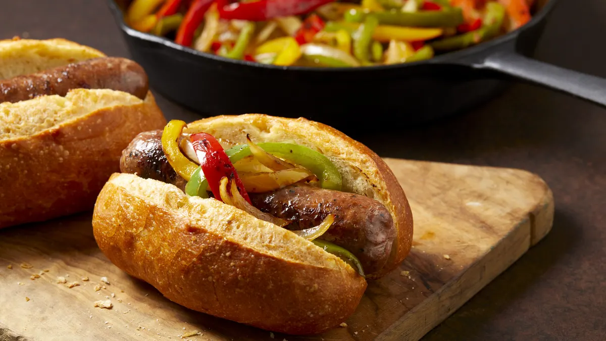 A small sausage and pepper sandwich on a roll sits on a wooden cutting board on a brown surface. A skillet of sauteed peppers is behind it.