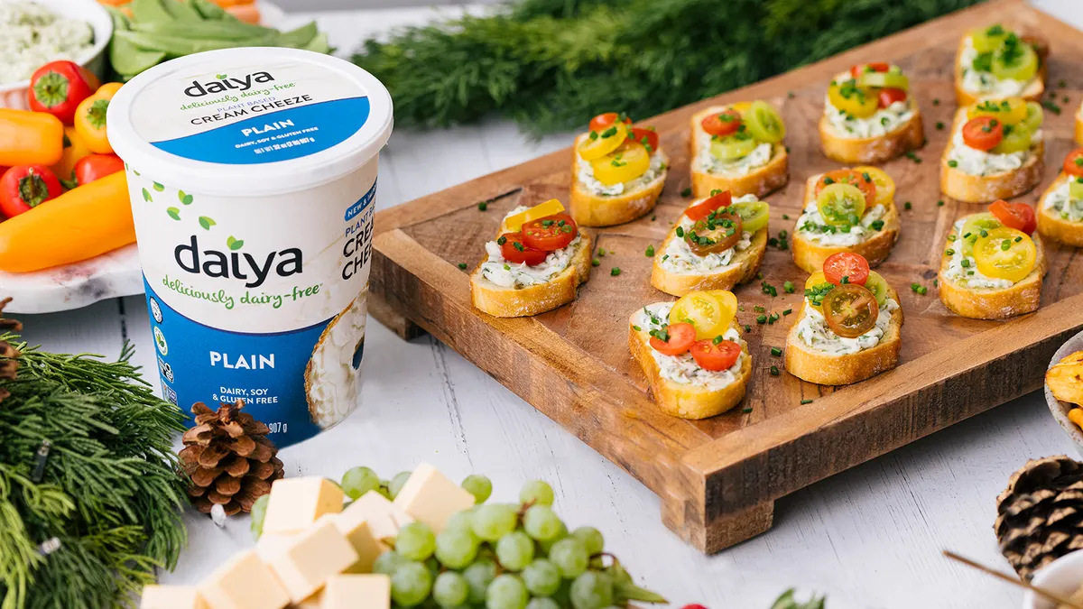A wooden serving board with small pieces of bread spread with cream cheese and topped with sliced cherry tomatoes and chives sits on a counter next to a tub of Daiya Cream Cheeze and other items.