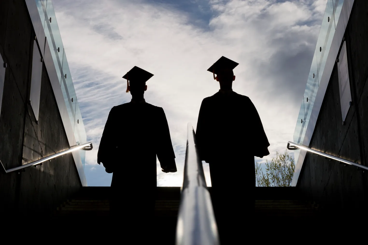 This is the silhouette of two college graduates in cap and gown climbing up a set of steps. This shot is backlit with bright morning sunshine in the background.