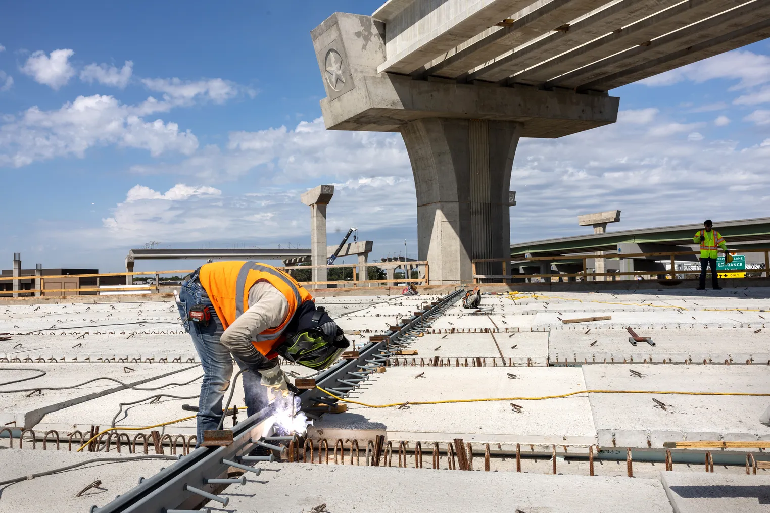 A person in a reflective vest and utility mask bends over their work, with unfinished concrete bridge parts in the background.