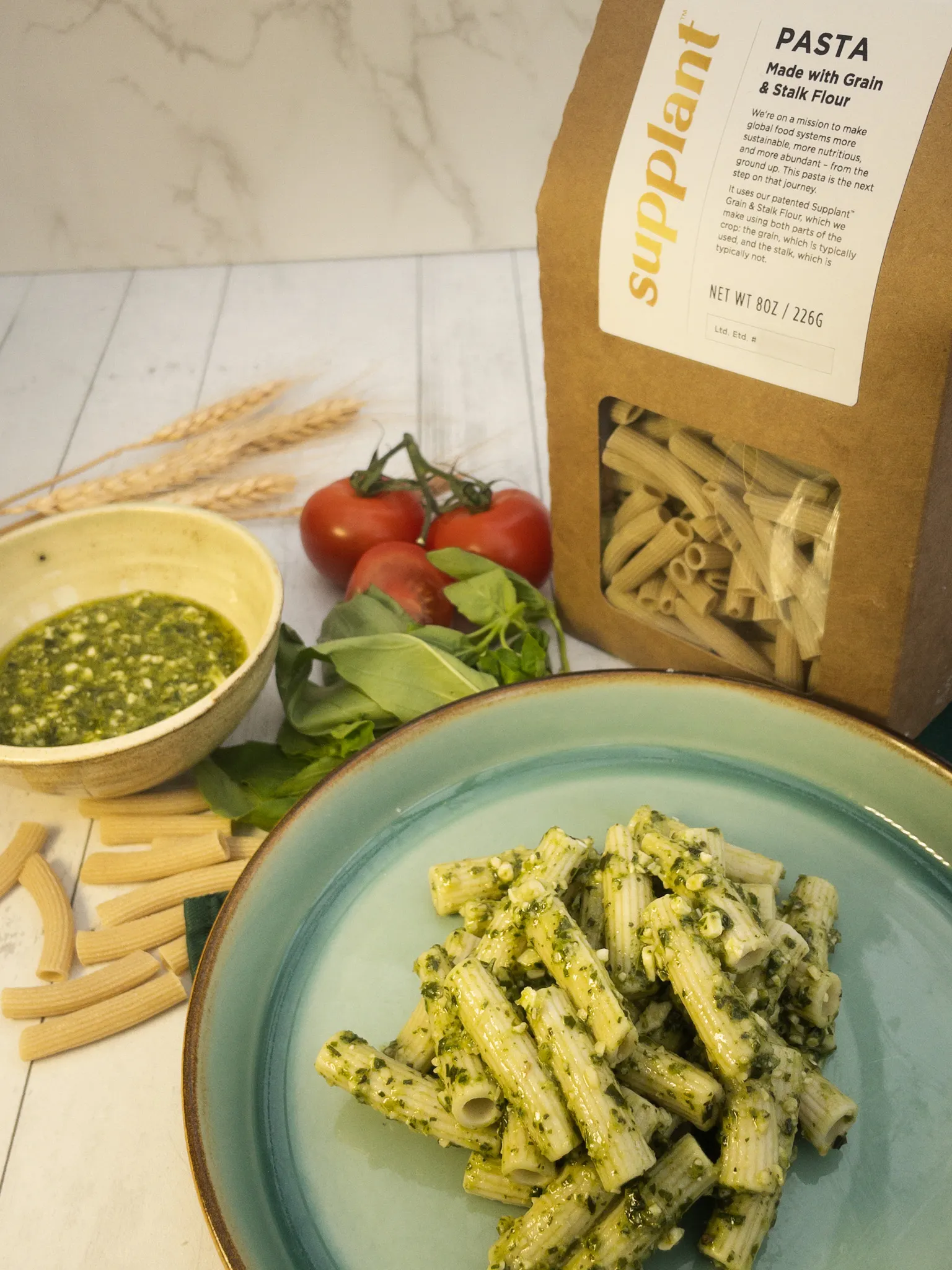 A light blue plate of pasta with pesto sauce is in the foreground, with a container of Supplant Pasta, a bowl of pesto sauce, noodles, basil leaves, tomatoes and wheat stalks behind it.