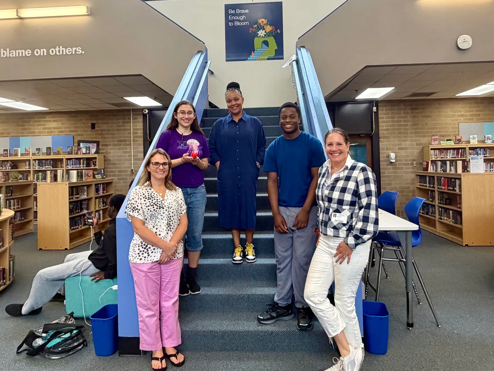 Five people stand on the stairs in a school library looking at the camera. They each stand on a different step on the stairs.