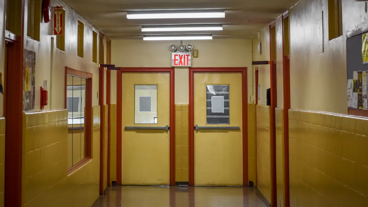 An empty school hallway looks straight at two doors. Over the doors is an exit sign.