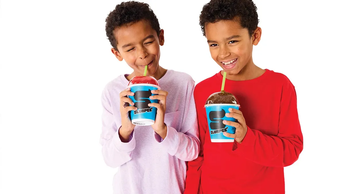 A photo of two children against a white background drinking beverages from disposable cups and smiling. The cups say Slurpee.