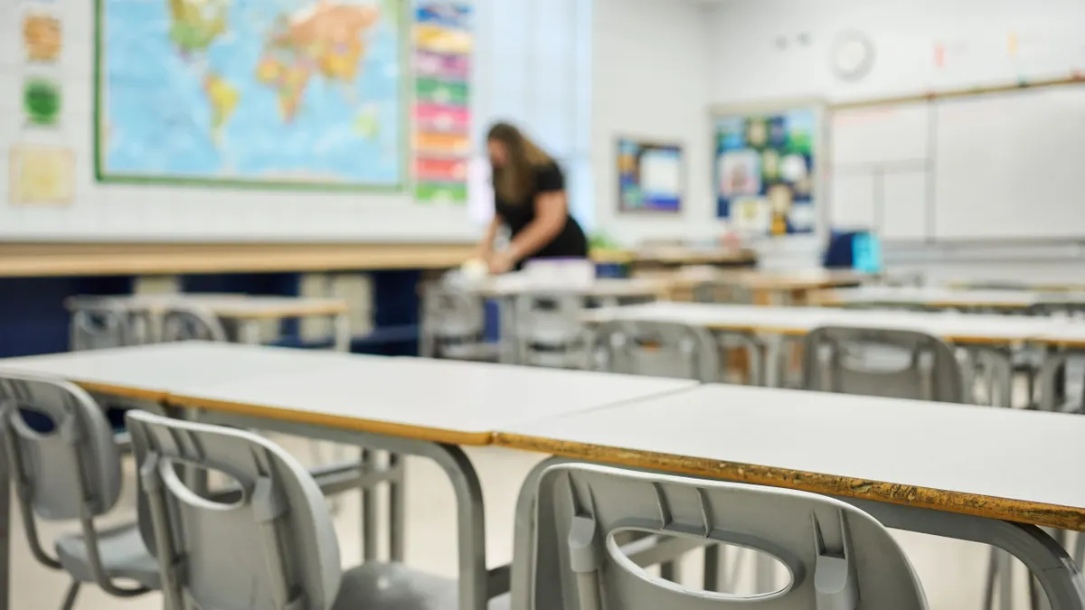 Chairs and desks sit in rows in a school classroom as a teacher works in the background.