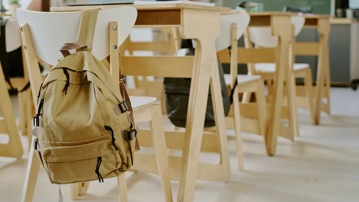 Desks and chairs are lined up in a classroom empty of students. A backpack hangs on one of the chairs.