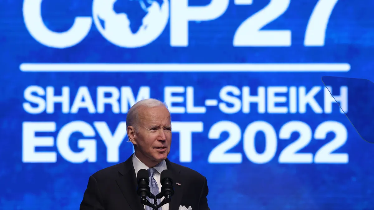 U.S. President Joe Biden stands on stage and speaks into a microphone at COP27 with the blue and white event logo and location on the screen behind him.