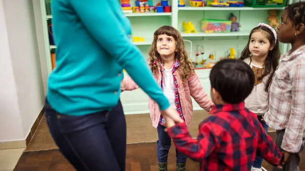 An adult stands in a circle with young children in a classroom. They all are holding hands.